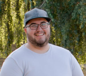 smiling man with broad shoulders, and dark beard, wearing light blue shirt, grey ball cap and glasses in front of natural greenery